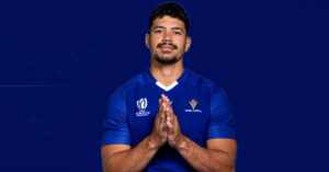 Samoan rugby player wearing a blue jersey with tournament and team logos holds his hands together against a dark blue background