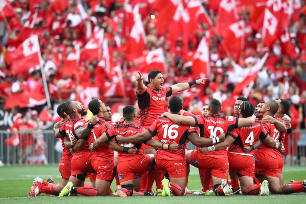 Tonga National Rugby League team performing the Sipi Tau war dance in front of a stadium of red flags.