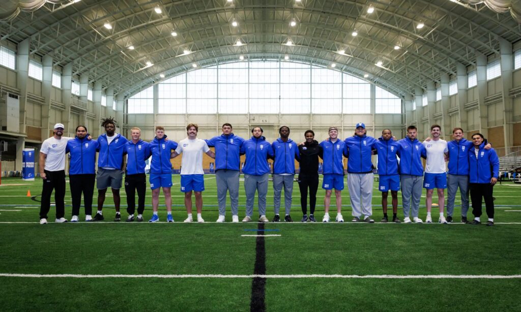 A wide-angle shot of a diverse group of college football players and coaches posing together on an indoor turf practice field during a Pro Day event.