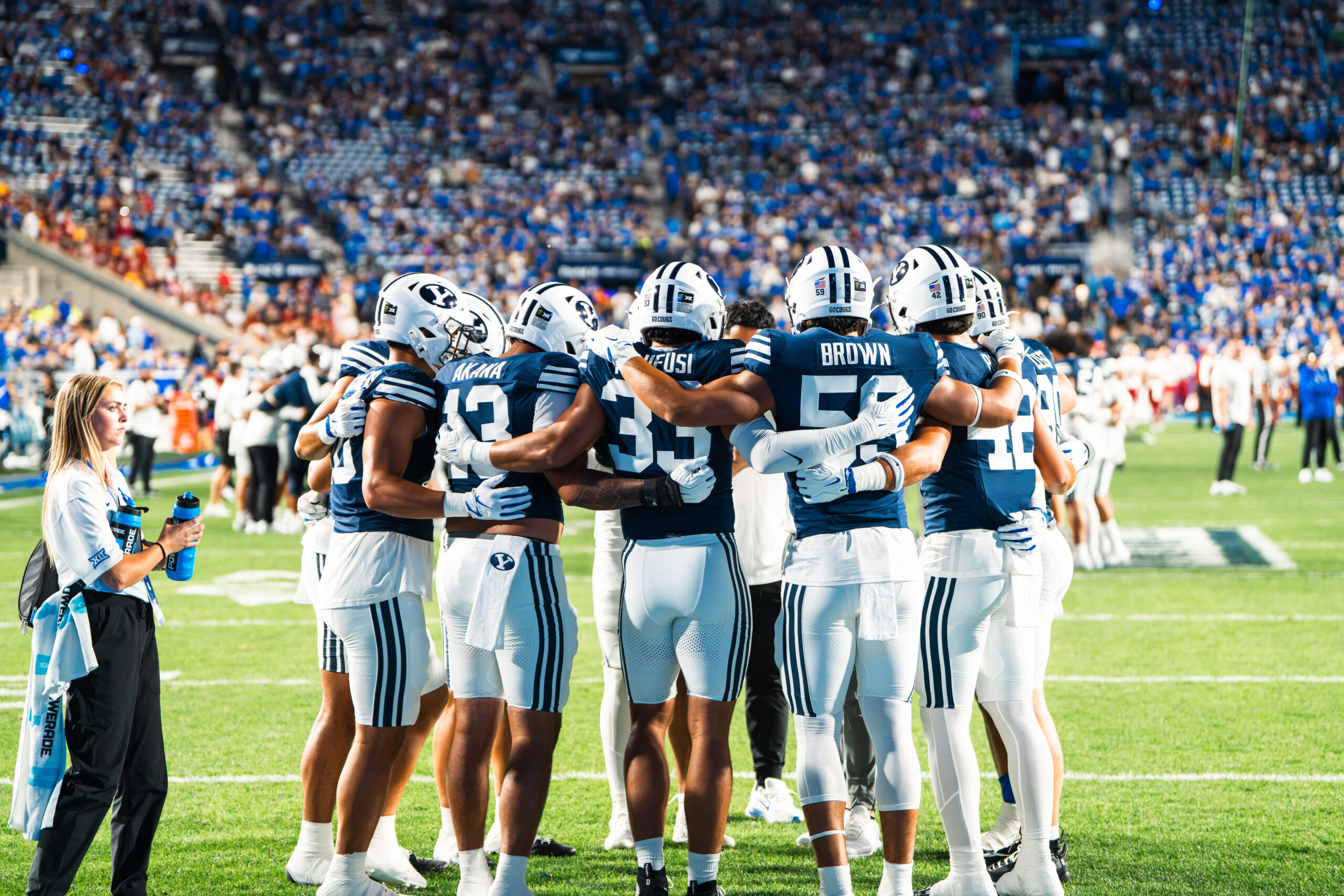 BYU football players gather in a pregame huddle on the field as a staff member watches nearby.